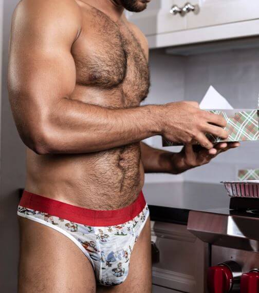 Man in kitchen wearing Sheer Thong St. Dick & Co with playful print and red waistband, showcasing comfort and style.
