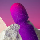 Glo-Girl Wand Massager Pink Purple close-up with droplets on a textured surface against a pastel background.