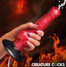 Creature Cocks Hell Hound Squirting Silicone Dildo held in hand with squirting feature, fiery background.