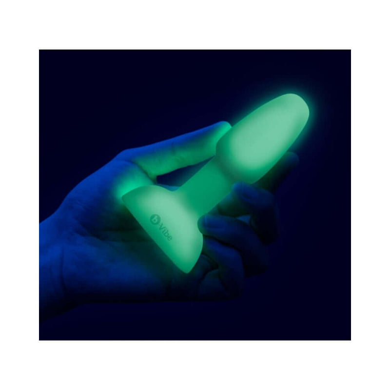 a hand holding a glowing b Vibe 10 Piece Asstronaut Glow in the Dark Butt Play Set toy in a dark setting