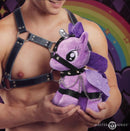 A man holding the Master Series Bondage Pony Purple plush toy in a faux leather harness.