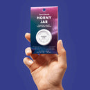 Bijoux Indiscrets Clitherapy Horny Jar Clitoral Balm 0.28 Oz packaging held in hand on a colorful background.