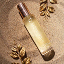 HighOnLove Dry Body Oil Spray bottle surrounded by decorative leaves on sand, showcasing a luxurious skin care product.