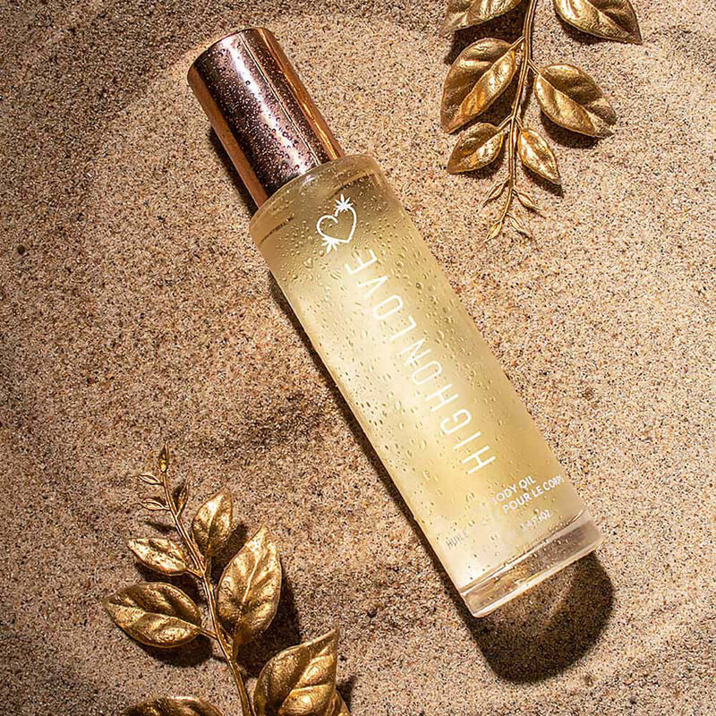 HighOnLove Dry Body Oil Spray bottle surrounded by decorative leaves on sand, showcasing a luxurious skin care product.