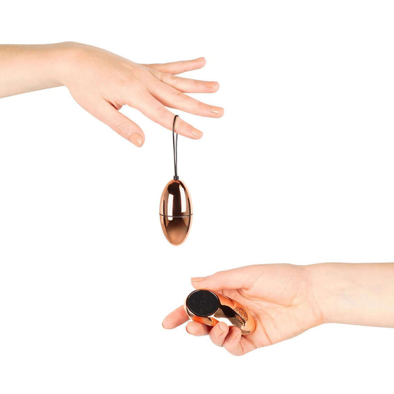 Two hands holding the Rosy Gold Nouveau Vibrating Egg and its remote control, showcasing elegance and playful control.