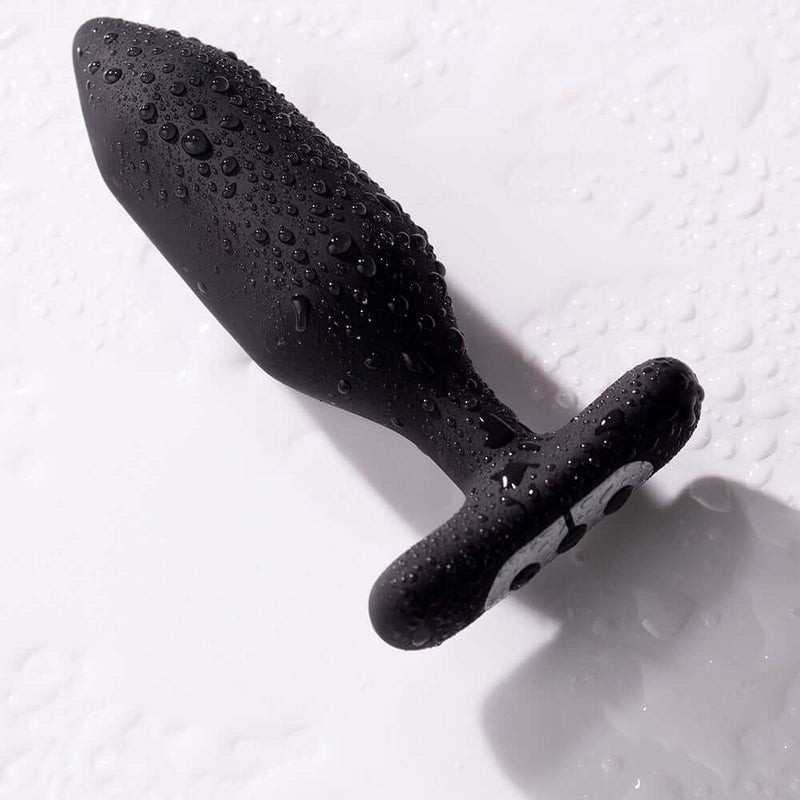 Je Joue Onyx Vibrating Butt Plug Black with water droplets on a surface, showcasing its smooth design.