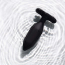 Je Joue Onyx Vibrating Butt Plug Black displayed on a smooth surface with gentle water ripples.