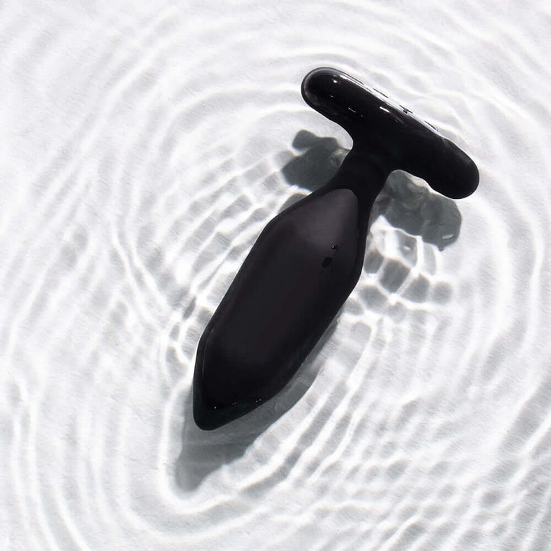 Je Joue Onyx Vibrating Butt Plug Black displayed on a smooth surface with gentle water ripples.