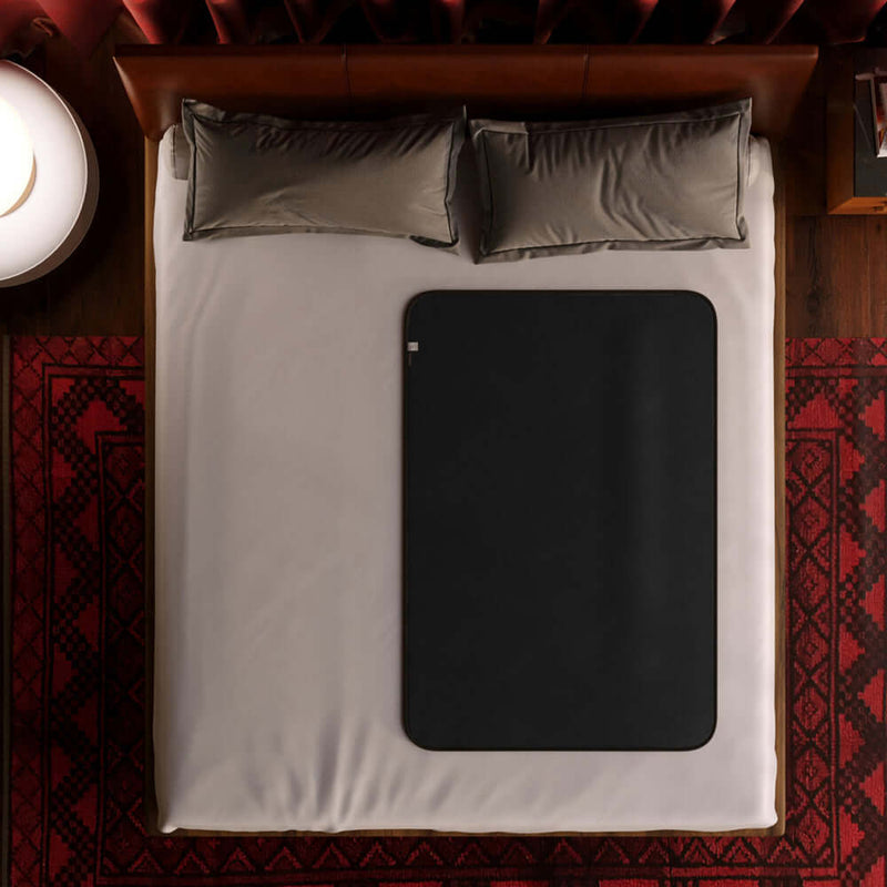 Liberator Fascinator Waterproof Throw Travel Blanket Black on a bed with gray pillows and a patterned rug.