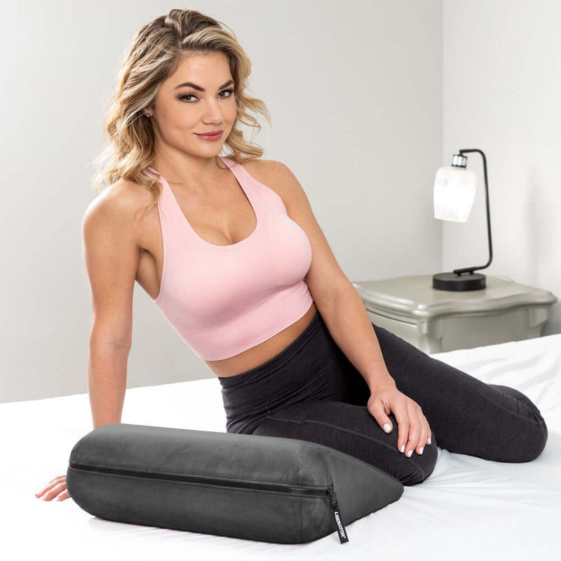 Liberator Jaz Original Sex Pillow Black positioned on a bed with a woman in a pink top, showcasing its discreet design.