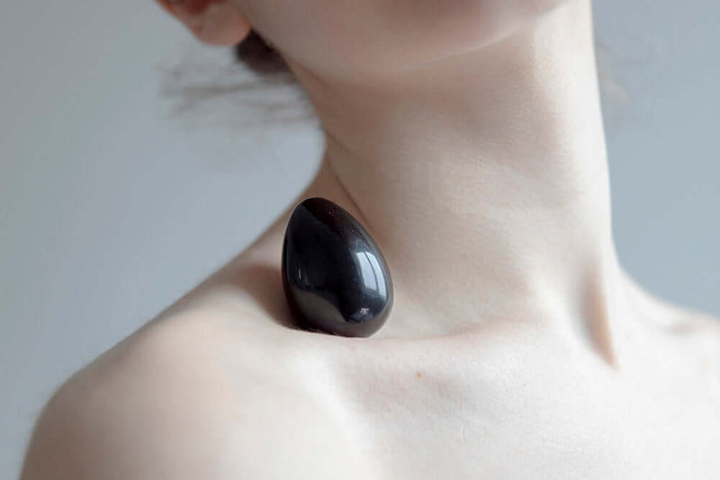 Chakrubs Xaga Yoni Egg Obsidian drilled resting on shoulder, showcasing its sleek, polished black obsidian surface.