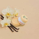 Sweetums Flavored Lip Balm Vanilla in a jar with vanilla flowers and beans, surrounded by water droplets.