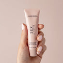 Hand holding Sex Au Naturel Vulva Balm Soothing Moisturizer tube against a soft pink background.