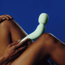 Dame Com Rechargeable Wand Vibrator Sage in hand resting on legs against a blue background.