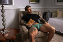 Male Power Eco Cozy Pouch Short Green M model relaxing in chair with book, showcasing comfort and fit.