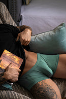 Male Power Eco Cozy Pouch Short Green M modeled by a man reading on a couch, showcasing comfort and style.