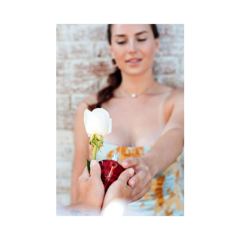 A woman in a floral dress receiving the Eye of Love Matchmaker Red Diamond Attract Her Parfum 1 Oz with a white rose.