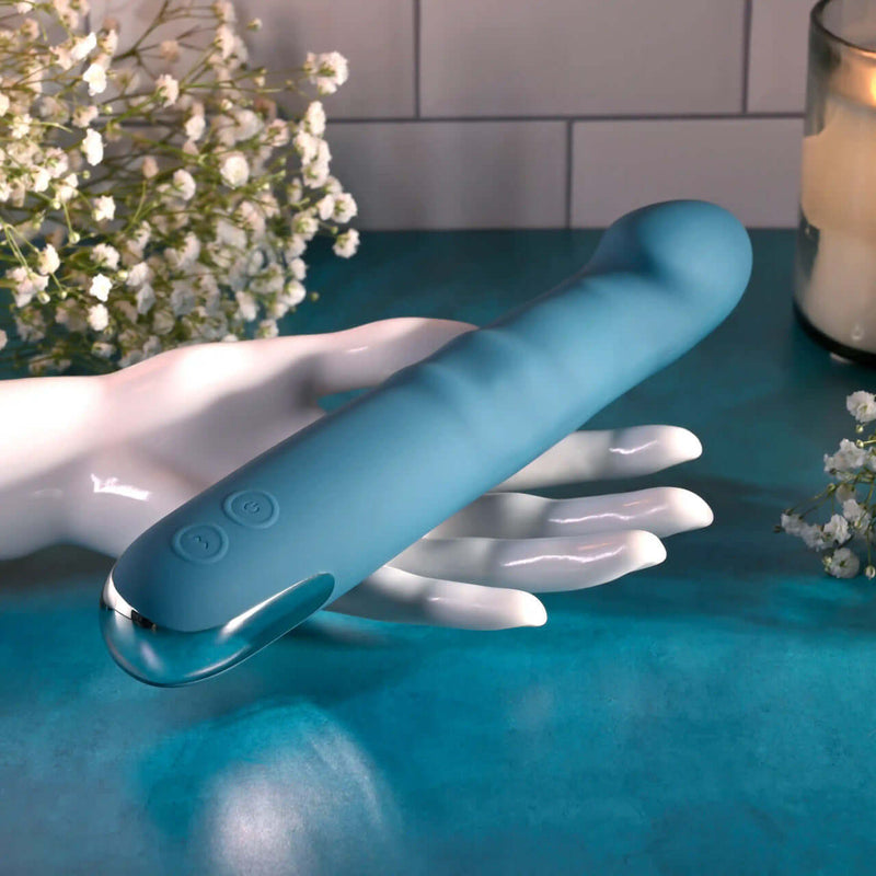 Rechargeable blue silicone dual vibrator resting on a hand, surrounded by delicate flowers and candlelight.