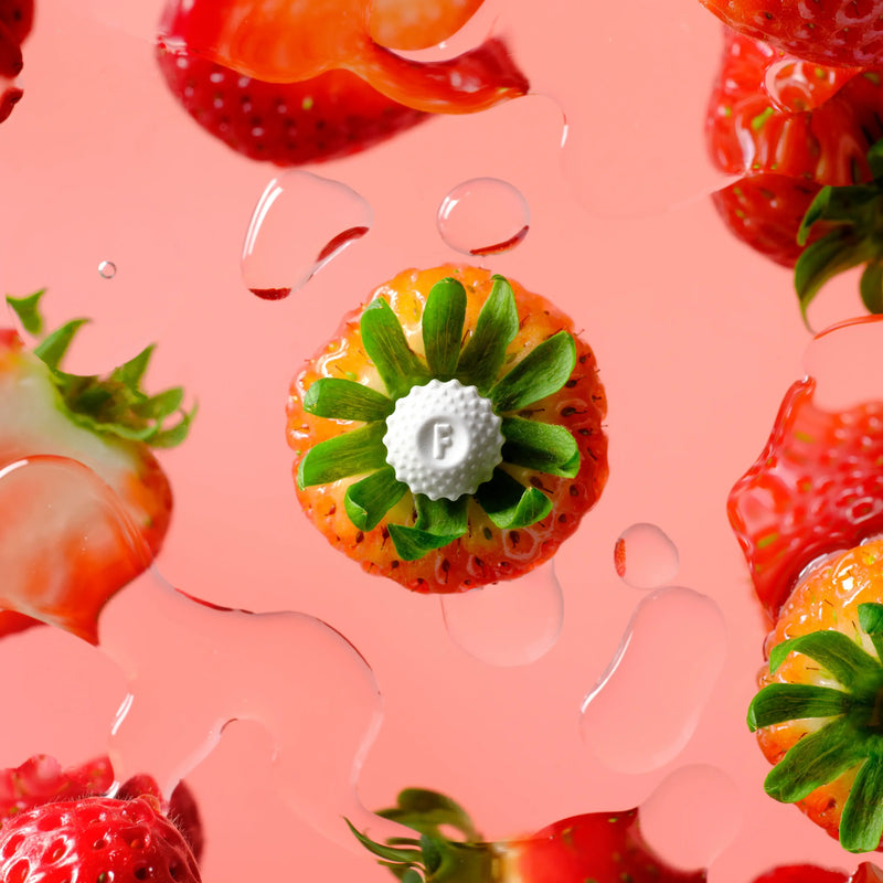 Fresh strawberries with a white F-shaped mint surrounded by juicy strawberry slices and droplets on a pink background.