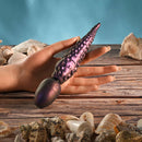 Gender X Into the Deep Silicone Vibrating Dildo Purple held in a hand, set on a background of seashells.