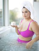 Woman in pink bikini holding Jimmyjane Natural Massage Oil Candle 4.5 Oz Santal while relaxing in a bathtub.