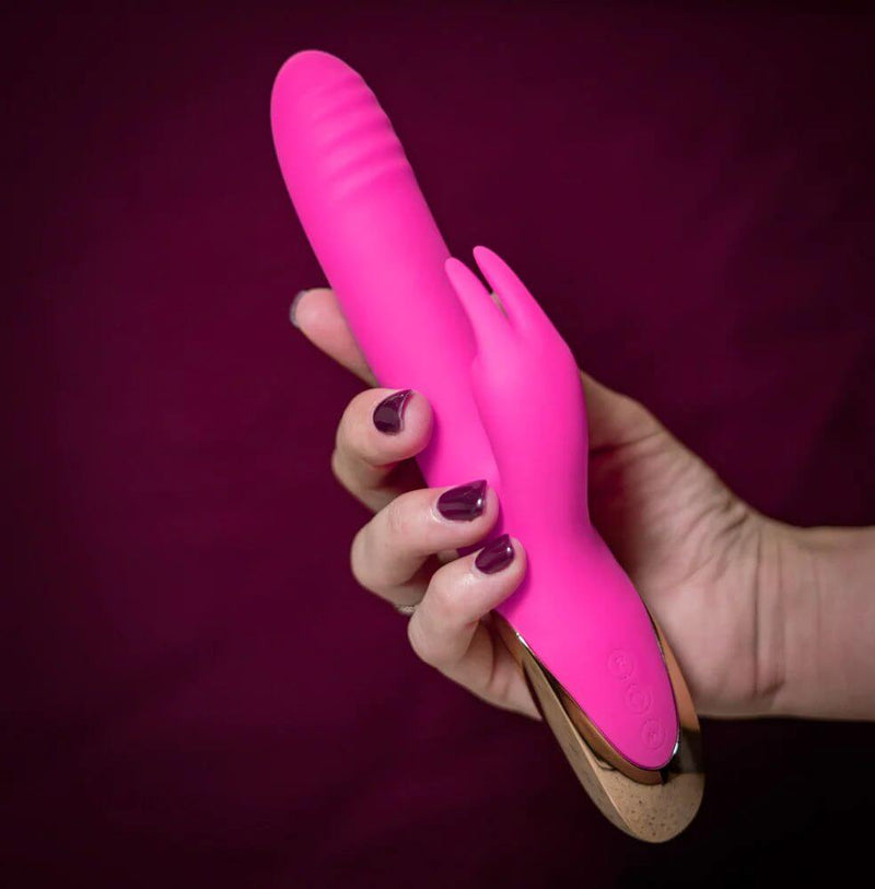 Hand holding pink silicone rabbit vibrator with clitoral stimulator and gold accents on a dark background.