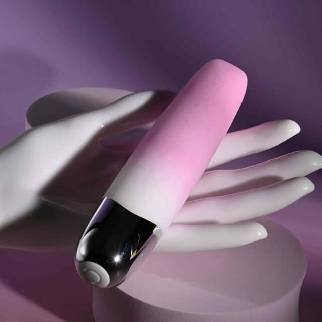 Pink rechargeable silicone vibrator with clitoral stimulator resting on a hand against a purple background.