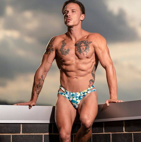 Man in patterned thong briefs with tattoos poses on rooftop, showcasing bold summer fashion in geometric swimwear.