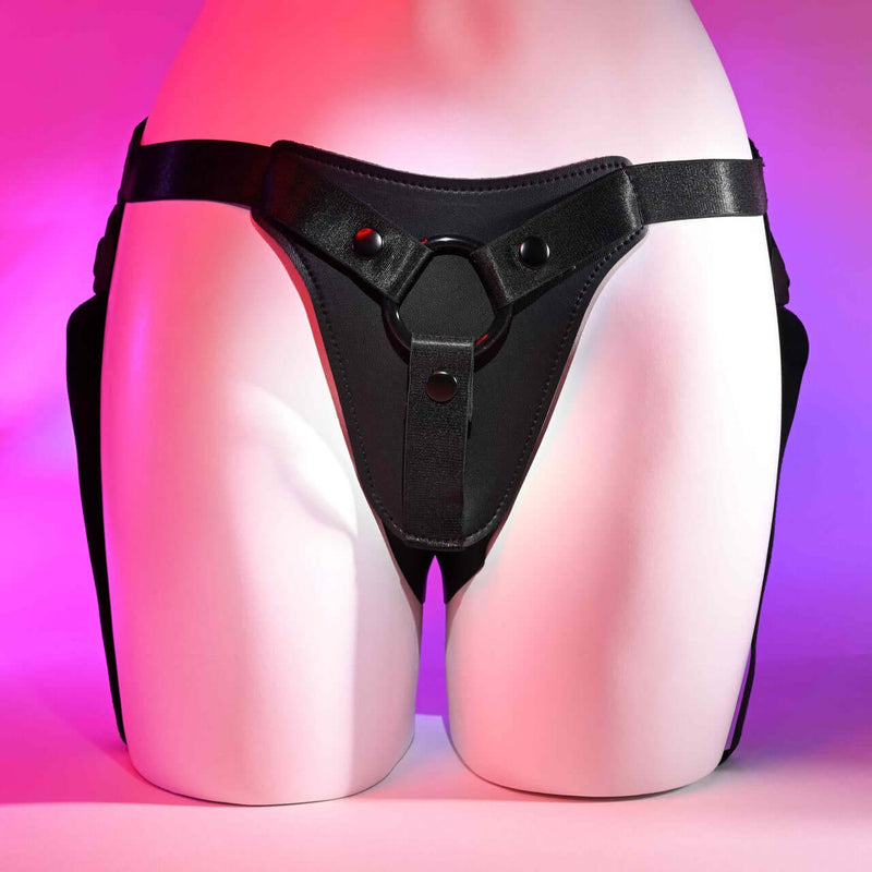 Fully adjustable faux-leather strap-on harness from Gender X, showcasing secure waist and leg straps on a mannequin with a pink background