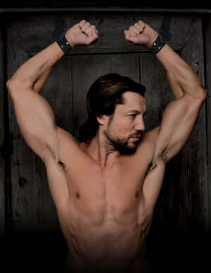Man with muscular build restrained in door cuffs, showcasing BDSM bondage setup with industrial strength straps.