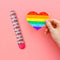 Pride Vibe Pink from Blush Novelties beside a hand holding a rainbow heart on a pink background