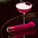 Oh My Gem Classy Garnet Red Vibrator next to an elegant cocktail garnished with cherries on a dark reflective surface.