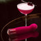 Oh My Gem Classy Garnet Red Vibrator next to an elegant cocktail garnished with cherries on a dark reflective surface.