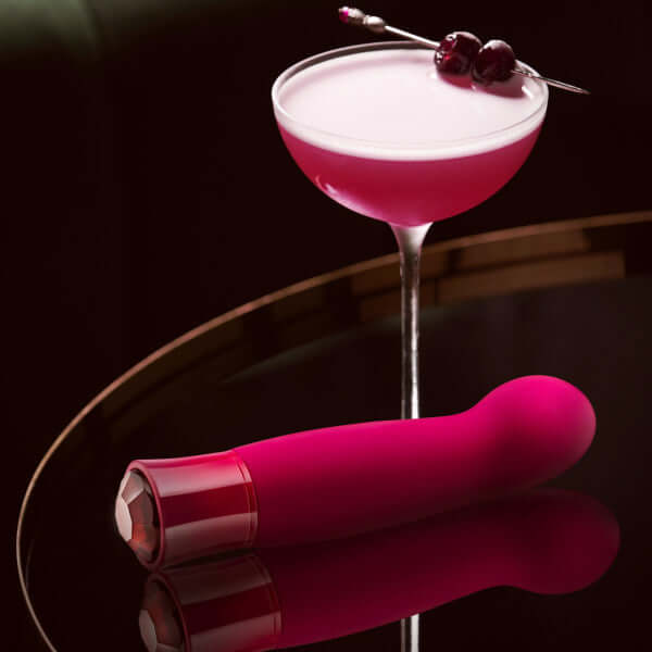 Oh My Gem Classy Garnet Red Vibrator next to an elegant cocktail garnished with cherries on a dark reflective surface.