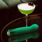 Oh My Gem Enchanting Emerald Vibrator next to a green cocktail on a reflective surface.