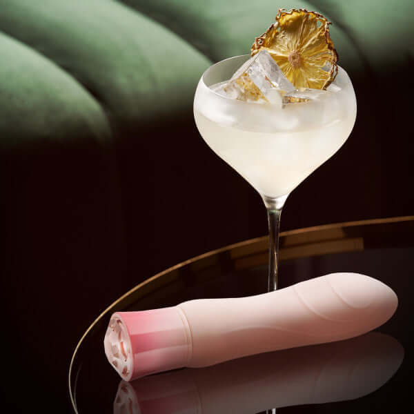 Oh My Gem Elegant Morganite Pink Vibrator by Blush Novelties alongside a cocktail garnish with dried pineapple on a reflective surface.