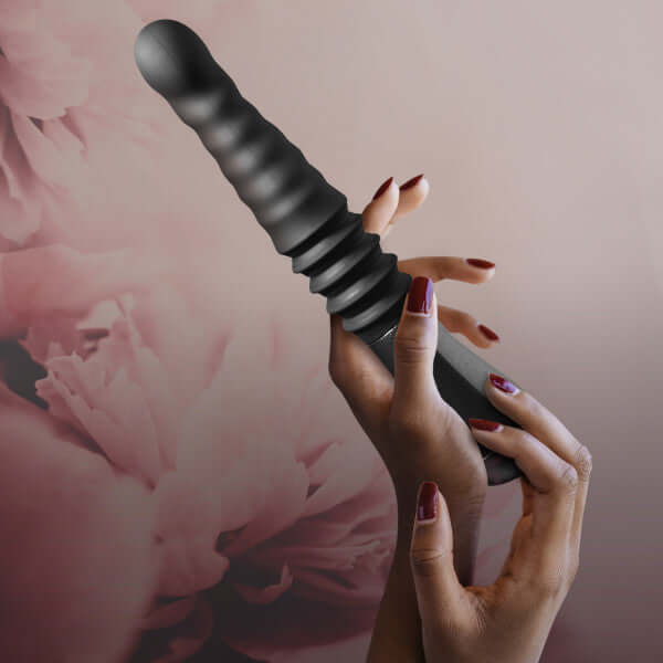 Person holding Temptasia Lazarus Black Thrusting Dildo with a ribbed shaft against a floral background.