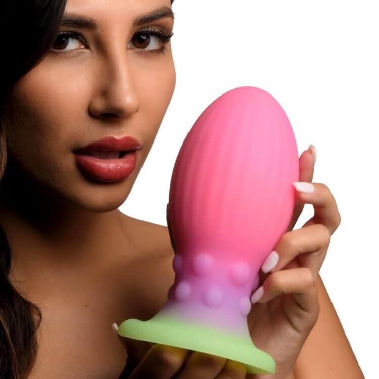 Woman holding Creature Cocks Xeno Glow In The Dark Silicone Egg, a pink and green alien-inspired toy for exploring fantasy desires