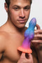 Man holding Creature Cocks Galactic Cock glow-in-the-dark alien dildo in colorful design with pink, orange, blue, and purple hues.